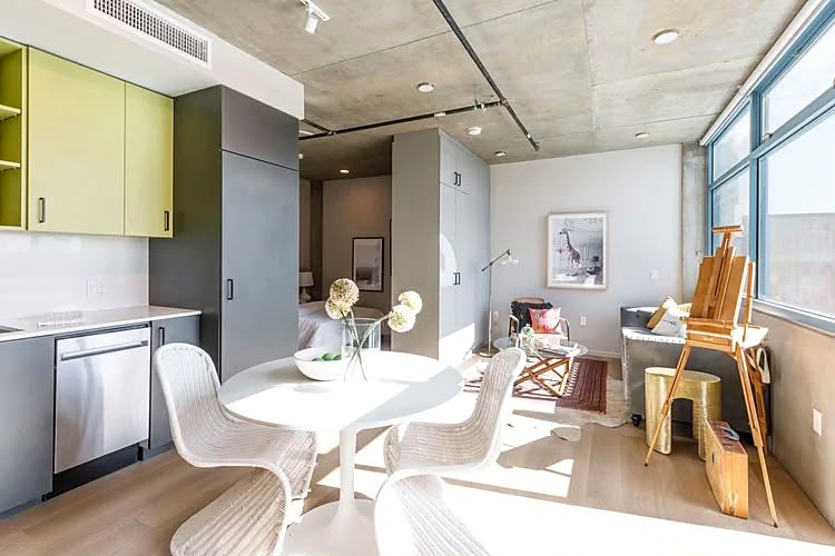 Open-plan studio with a green upper-cabinet kitchen along one wall and a dining table under the window.
